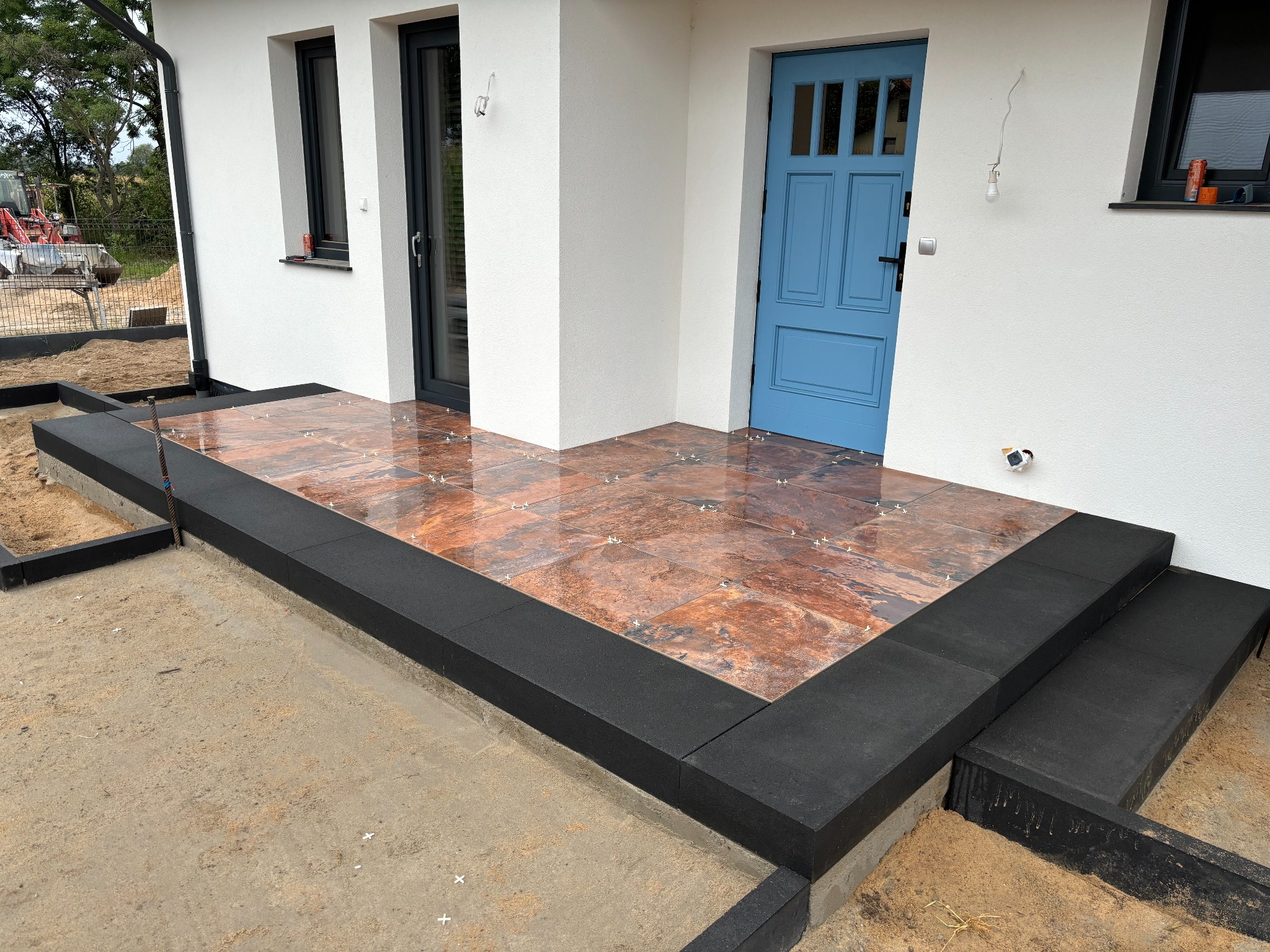 Nowo ułożona, błyszcząca, brązowo-pomarańczowa terakota na tarasie przed wejściem do domu z błękitnymi drzwiami. Widoczne czarne obrzeża tarasu i przygotowane podłoże z piasku. W tle traktor.