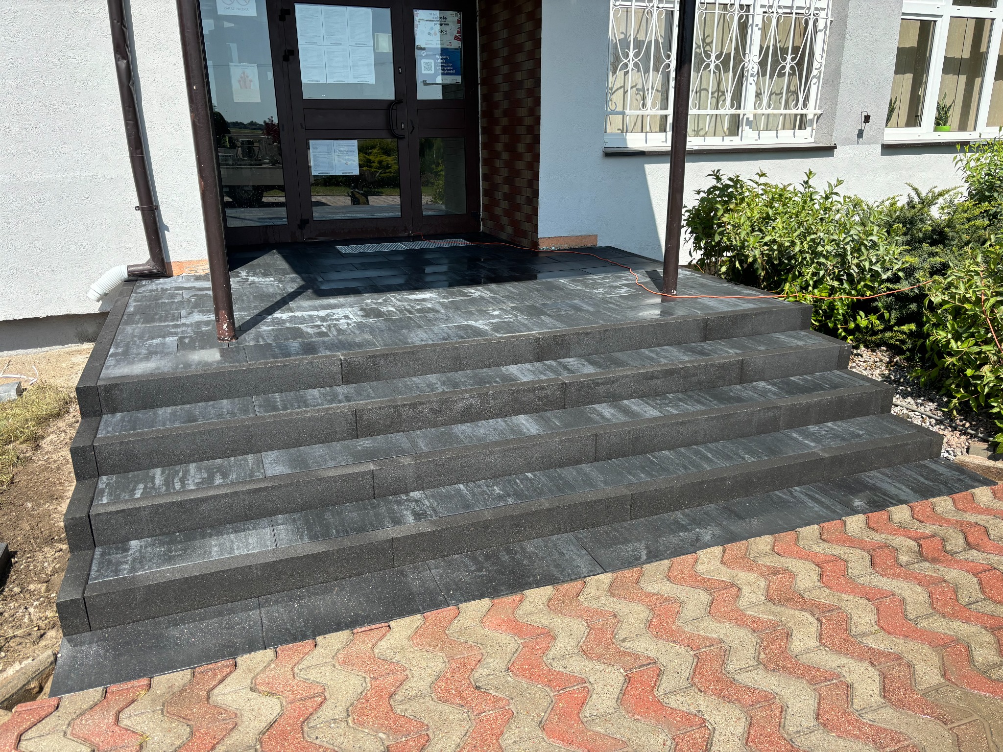Nowoczesne, czterostopniowe schody wejściowe wyłożone szarą kostką brukową, prowadzące do budynku z przeszklonymi drzwiami, kontrastujące z wzorzystą, czerwoną kostką na chodniku.