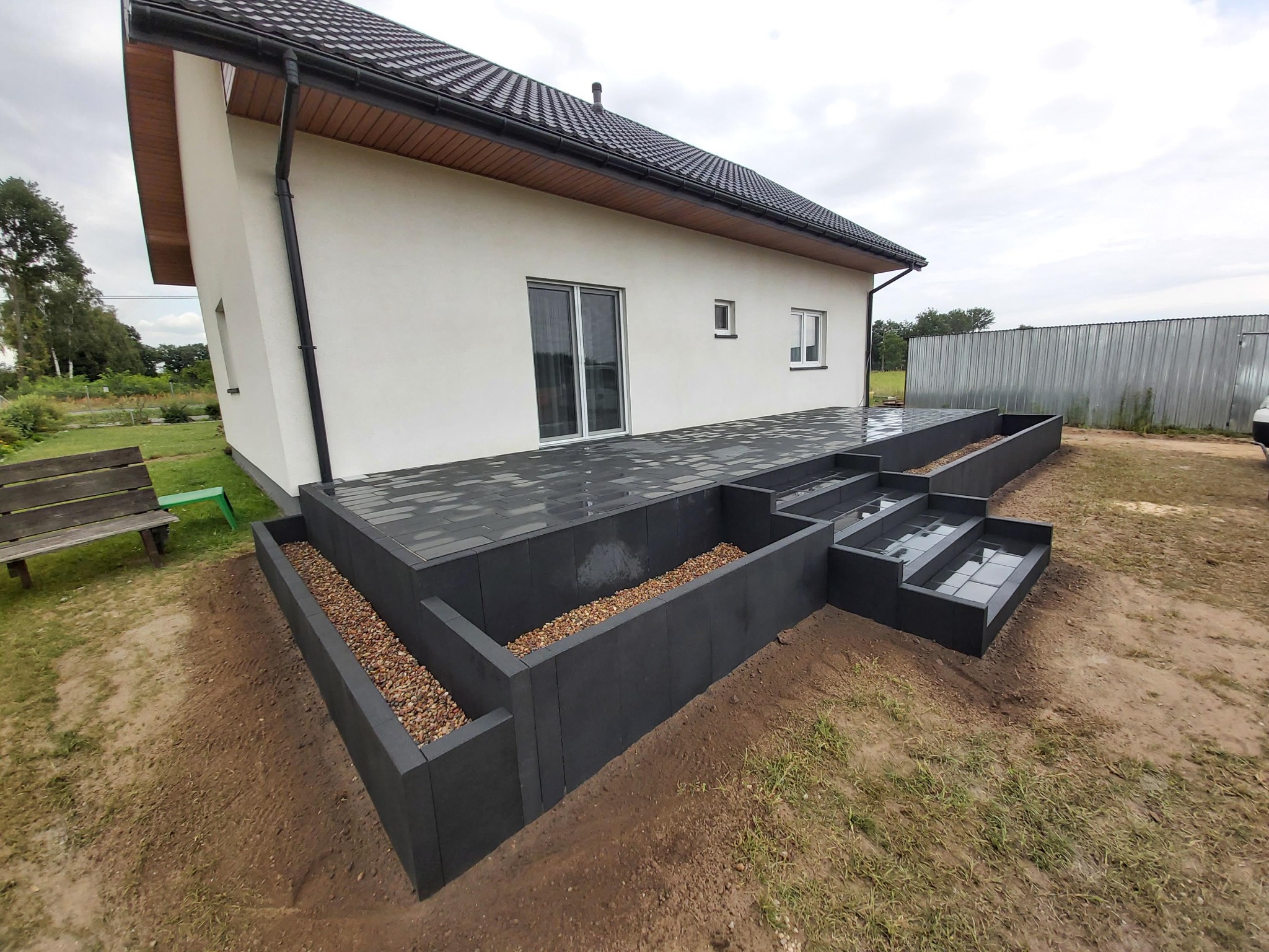 Nowoczesny taras z ciemnej kostki brukowej, schody i podwyższone rabaty z kamieniami ozdobnymi, przylegający do białego domu z szarą dachówką.