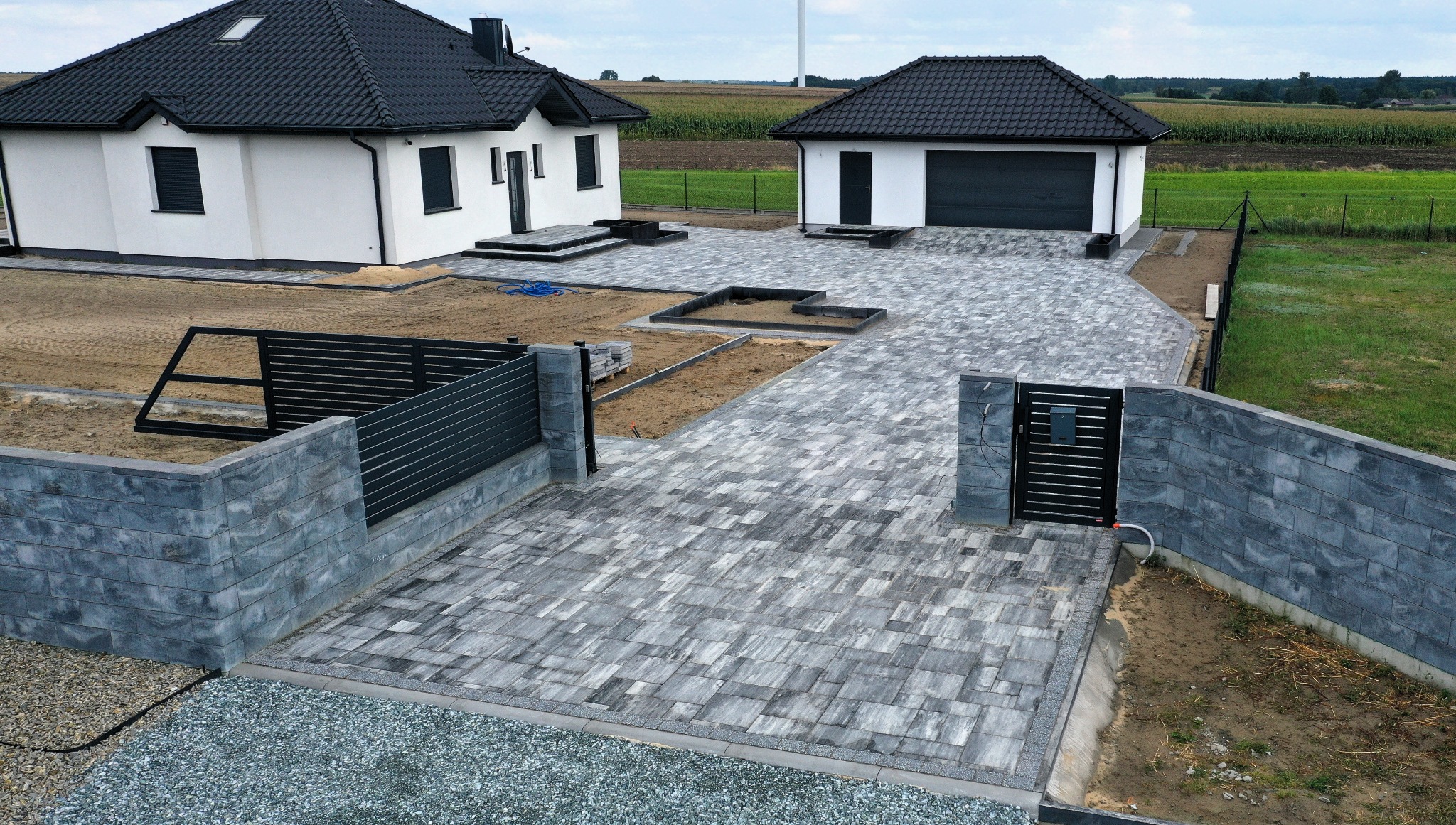 Nowoczesna posesja z szarej kostki brukowej, otoczona niskim, grafitowym ogrodzeniem z elementami betonowymi. Widok z góry na dom i garaż w tle.