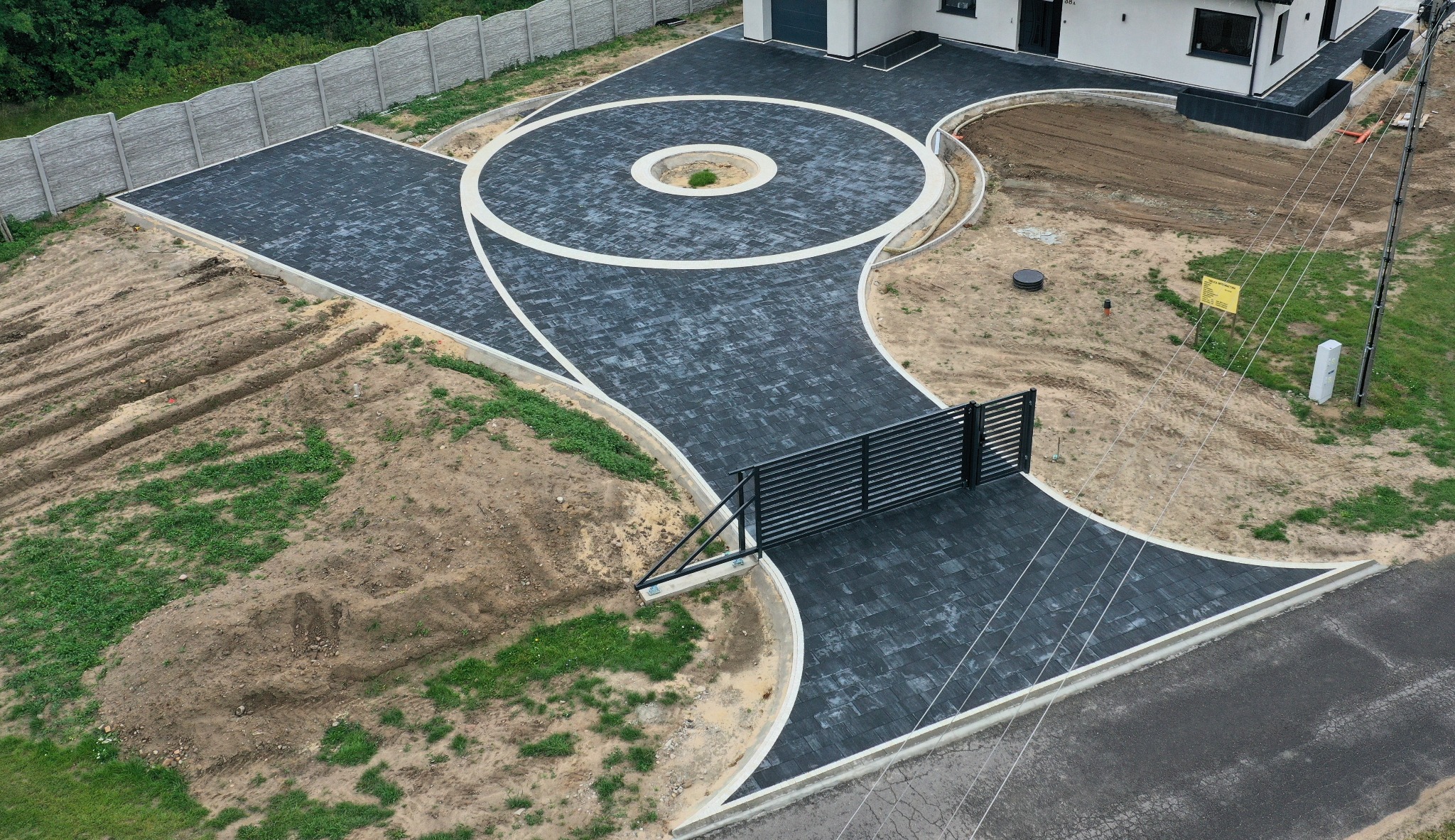 Nowoczesny podjazd z ciemnej kostki brukowej, z okrągłym wzorem i białymi obrzeżami. Widok z góry, częściowo zagospodarowany teren, brama wjazdowa.