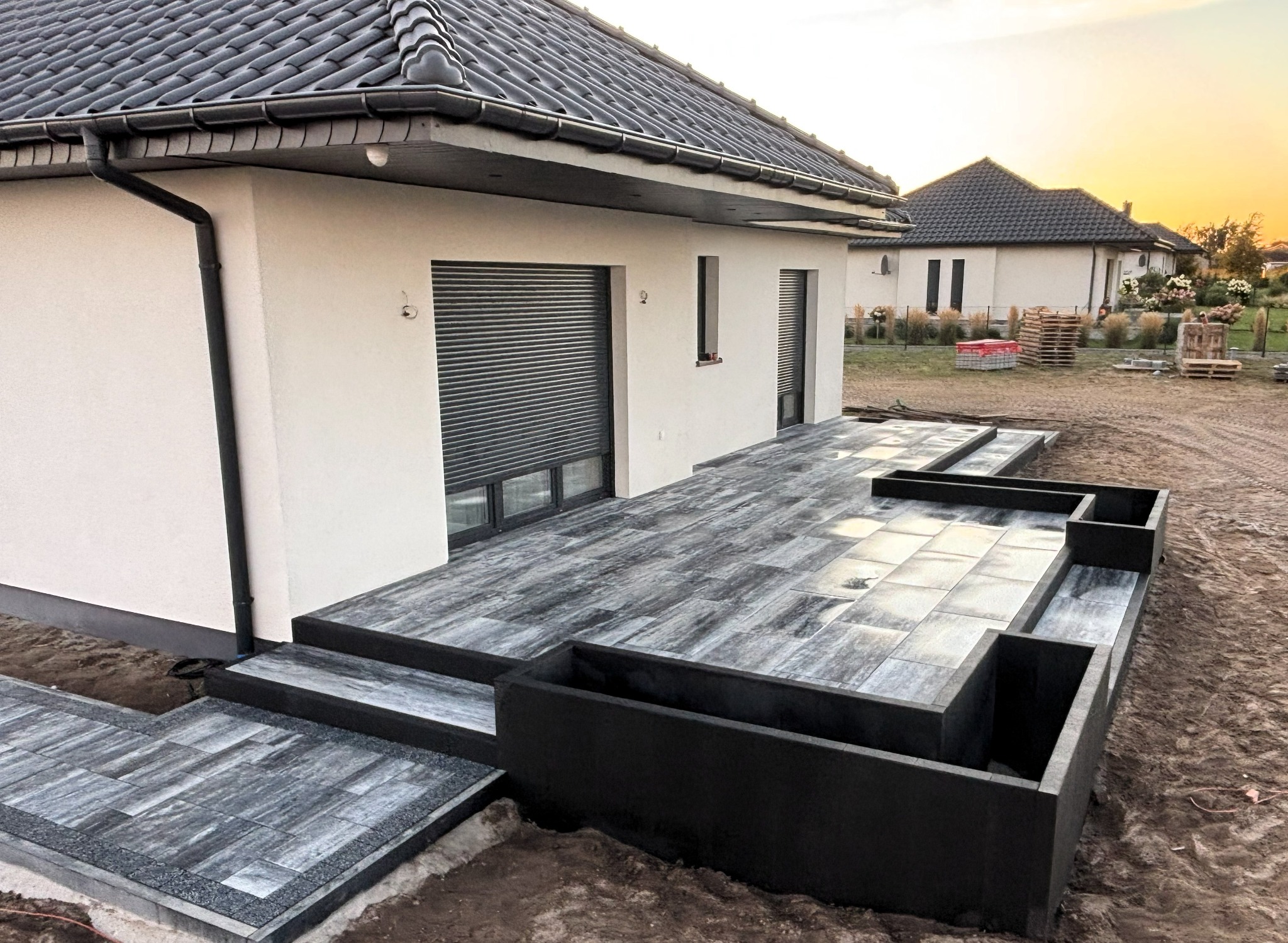 Nowoczesny taras z szarej kostki brukowej, zintegrowane donice, minimalistyczny design, dom jednorodzinny w tle, ciepłe światło zachodzącego słońca.