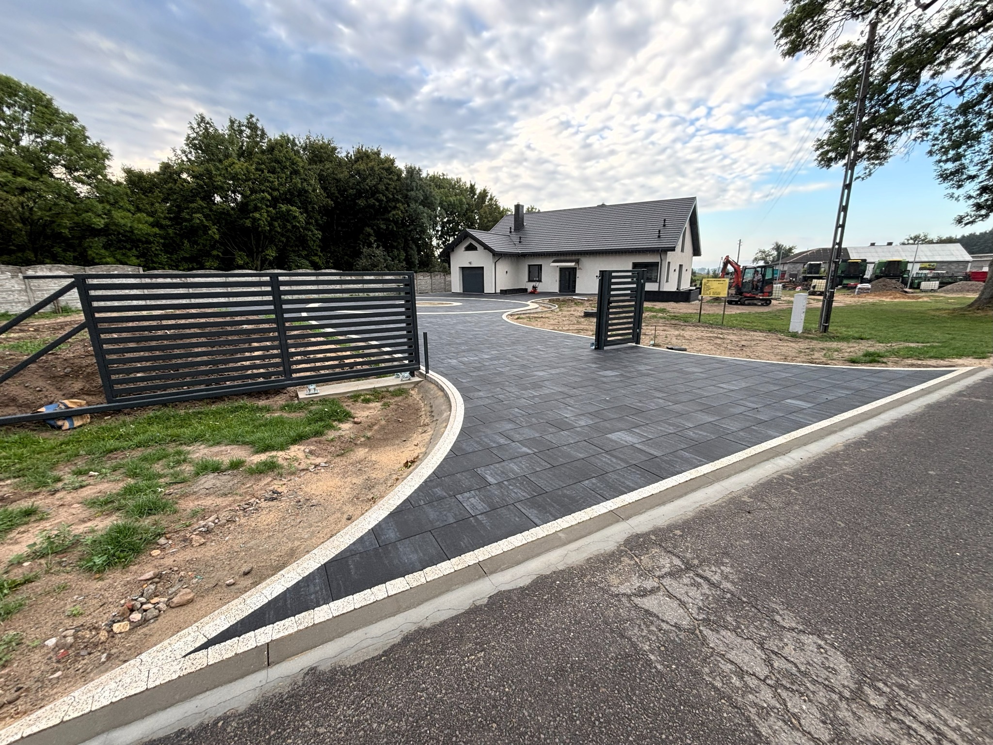 Nowoczesny podjazd z grafitowej kostki brukowej, obrzeża z jasnej kostki. W tle dom z szarą dachówką i czarnymi detalami, brama wjazdowa i częściowo widoczne ogrodzenie.