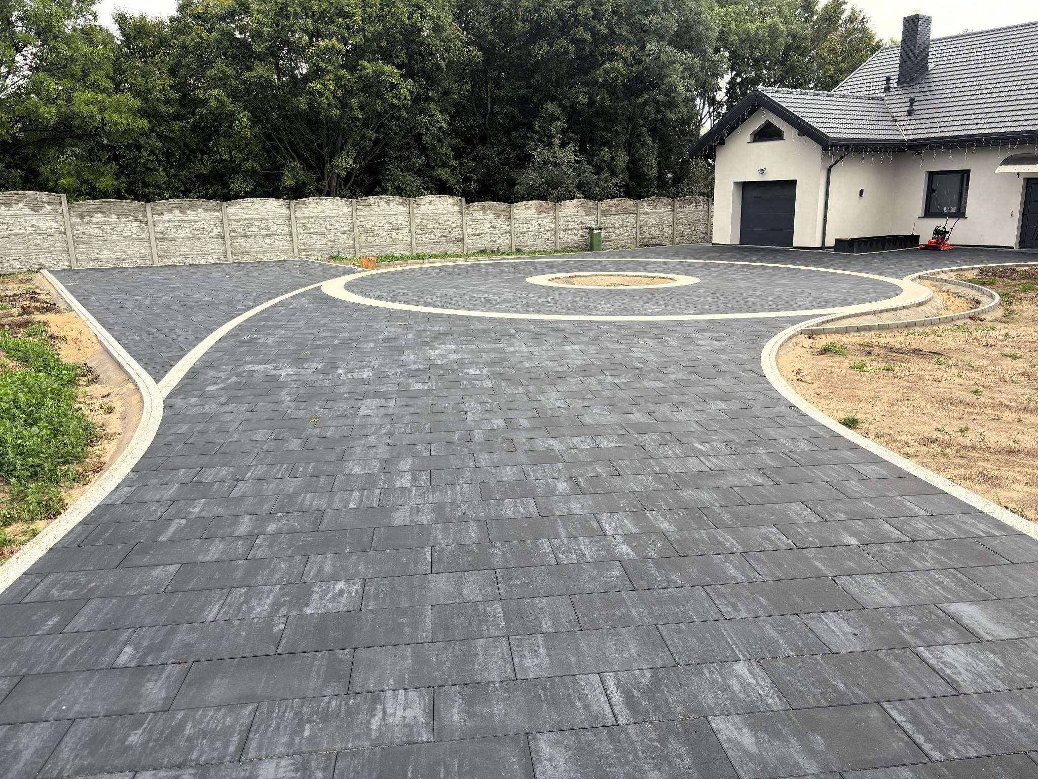 Ukończony podjazd z ciemnej kostki brukowej, z białymi obrzeżami, tworzący okrąg wokół trawnika. W tle ogrodzenie panelowe i budynek z garażem.