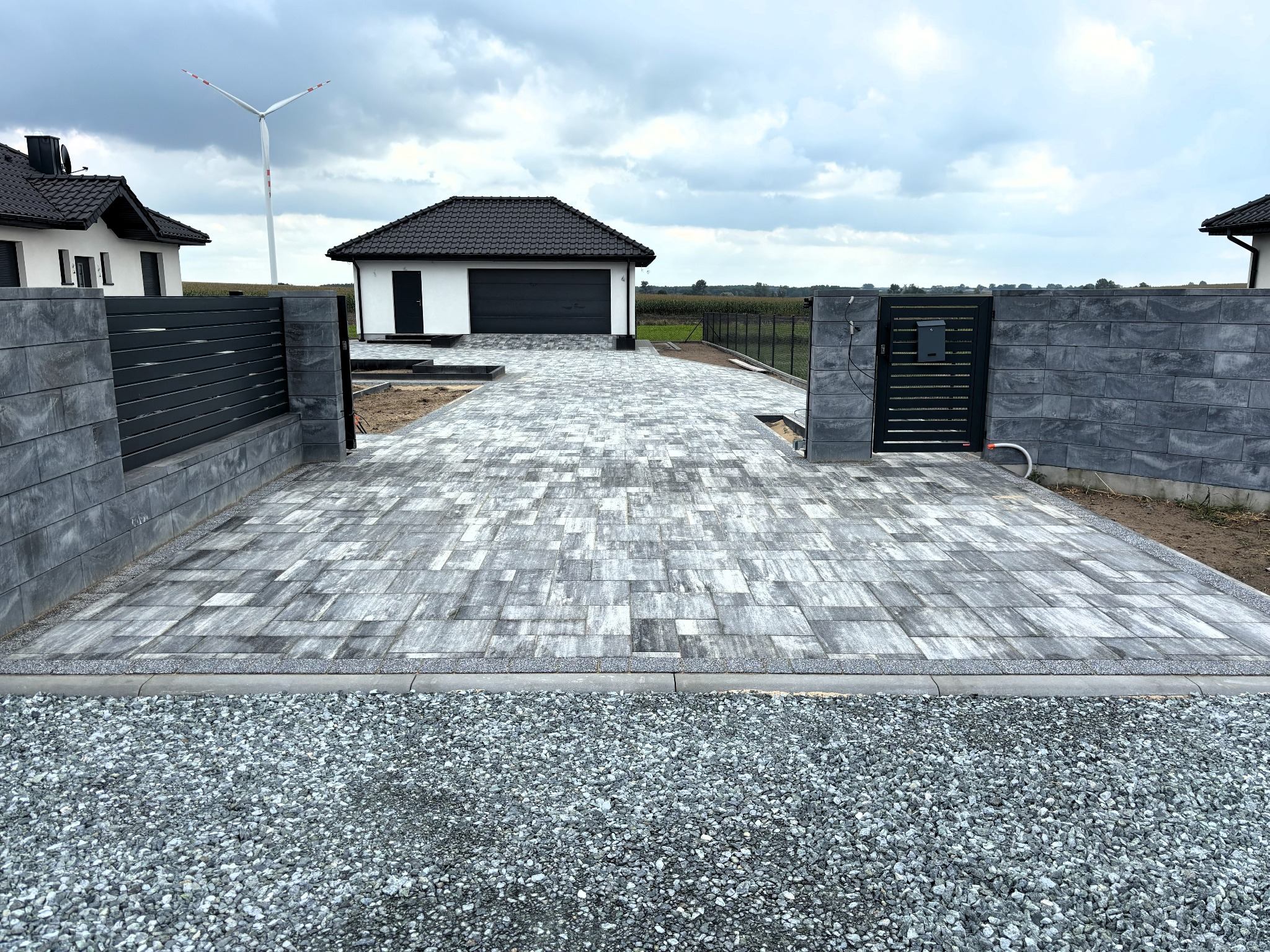 Ułożona kostka brukowa w Koło, prowadząca do garażu i furtki. Widoczny wiatrak w tle. Kostka w odcieniach szarości, ułożona we wzór. Nowoczesne ogrodzenie.