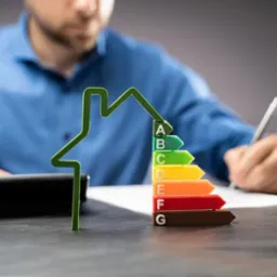 Mężczyzna wypełnia dokumenty obok modelu domu z kolorową skalą energetyczną A-G, kalkulator obok. Koncepcja świadectwa energetycznego.