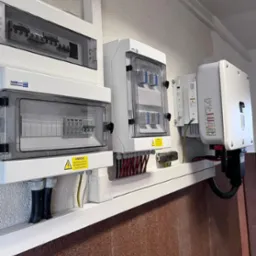 Rozdzielnice elektryczne i falownik fotowoltaiczny zamontowane na ścianie, widoczne przewody i ostrzeżenia o napięciu.