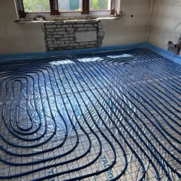 F.H.U Taurus Tadeusz Kuśpiel - Niebieskie rury ogrzewania podłogowego ułożone na srebrnej folii refleksyjnej w surowym wnętrzu z oknem, widoczne fragmenty cegieł i instalacji elektrycznej.