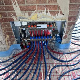 F.H.U Taurus Tadeusz Kuśpiel - Rozdzielacz ogrzewania podłogowego z niebiesko-czerwonymi rurami na folii refleksyjnej, zamontowany w surowej wnęce z cegły.