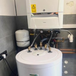 F.H.U Taurus Tadeusz Kuśpiel - Biały piec gazowy De Dietrich zamontowany nad zasobnikiem ciepłej wody, widoczne podłączenia hydrauliczne i elektryczne, ściana z szarą glazurą.