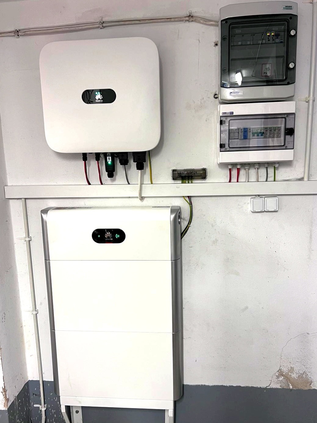 Biały inwerter fotowoltaiczny i akumulator energii na ścianie z szarą lamperią, widoczne skrzynki elektryczne i listwy z przewodami.