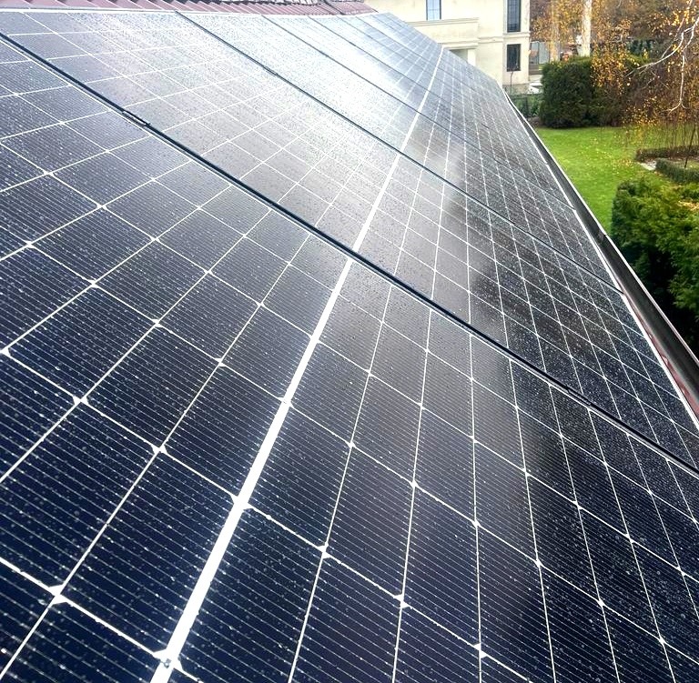 Panel słoneczny na dachu domu w deszczowy dzień, widoczne krople wody na powierzchni modułów fotowoltaicznych, w tle zielony trawnik i drzewa.