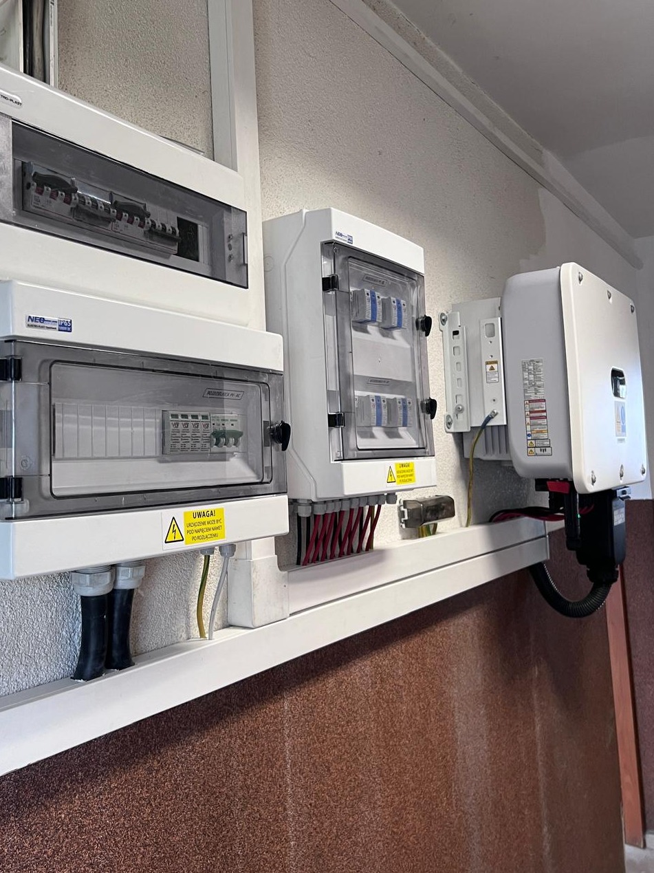 Rozdzielnice elektryczne i falownik fotowoltaiczny zamontowane na ścianie, widoczne przewody i ostrzeżenia o napięciu.