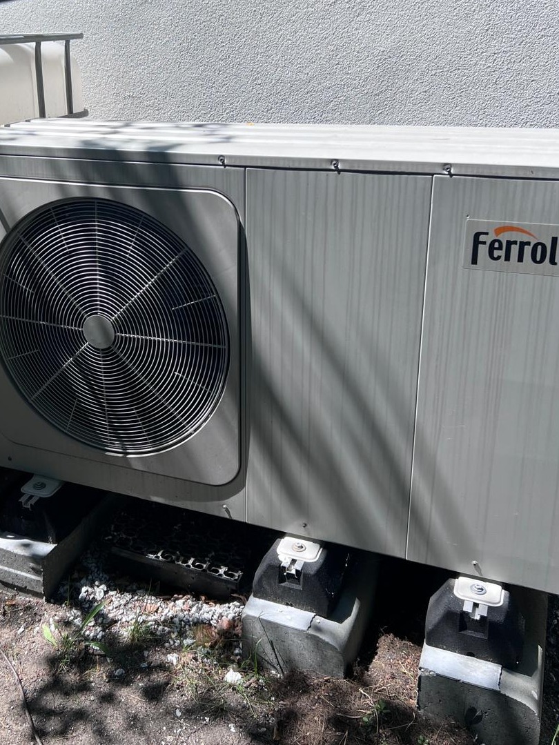 Zewnętrzna jednostka pompy ciepła Ferroli, zamontowana na betonowych podstawach, widoczne elementy instalacji i logo producenta.