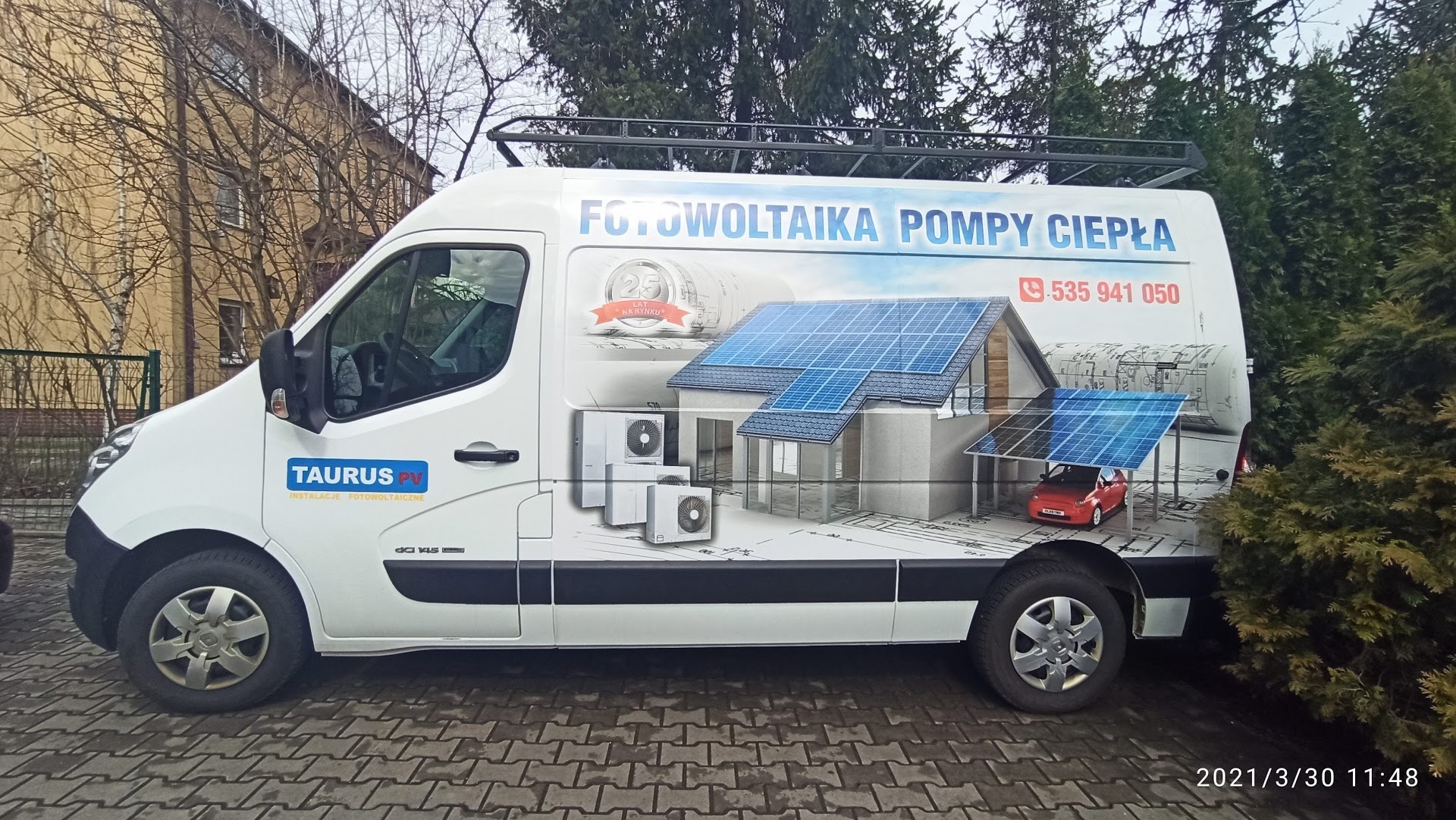 Biały samochód dostawczy z grafiką przedstawiającą dom z panelami słonecznymi, pompy ciepła i logo firmy Taurus PV, zaparkowany na brukowanej powierzchni.