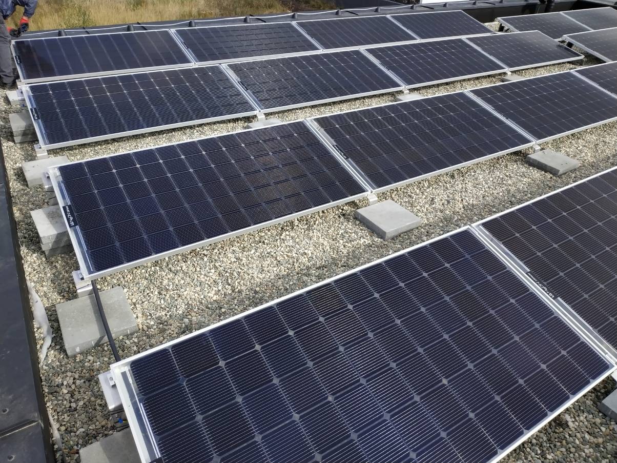 Rząd paneli słonecznych na płaskim dachu pokrytym żwirem, wspartych na betonowych blokach, widok z góry pod kątem.