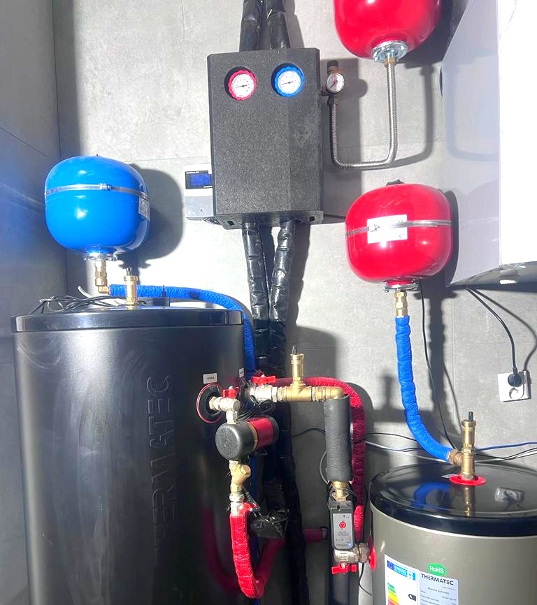 Dwa zbiorniki akumulacyjne Thermatec z niebieskimi i czerwonymi naczyniami przeponowymi, połączone rurami w izolacji, zamontowane przy szarej ścianie, z widoczną czarną skrzynką sterującą...