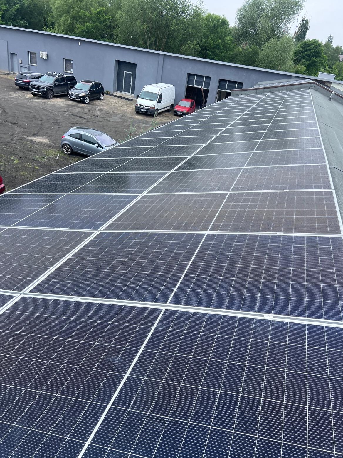 Rząd paneli słonecznych na dachu budynku przemysłowego, widoczne samochody na parkingu w tle, pochmurne niebo.