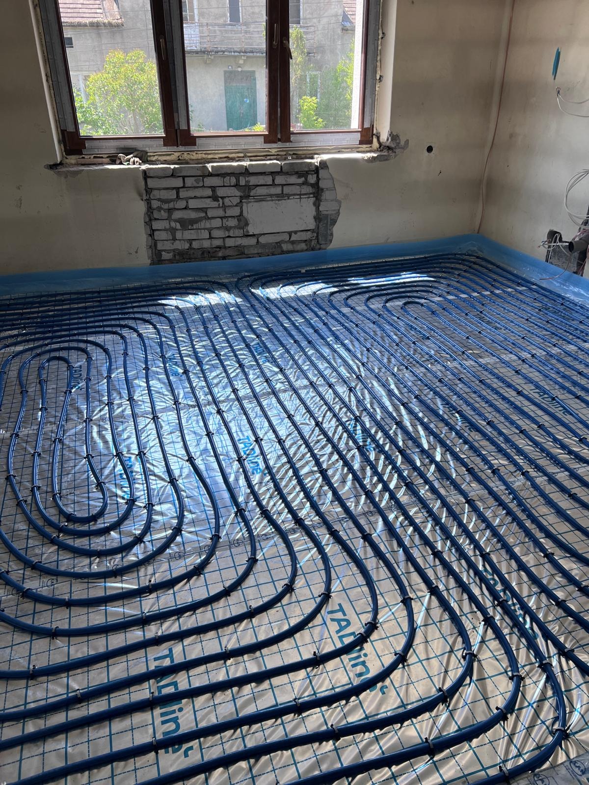 Niebieskie rury ogrzewania podłogowego ułożone na srebrnej folii refleksyjnej w surowym wnętrzu z oknem, widoczne fragmenty cegieł i instalacji elektrycznej.