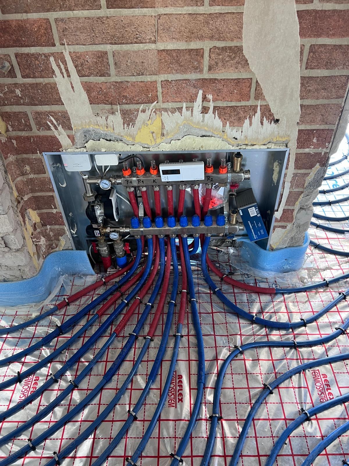 Rozdzielacz ogrzewania podłogowego z niebiesko-czerwonymi rurami na folii refleksyjnej, zamontowany w surowej wnęce z cegły.