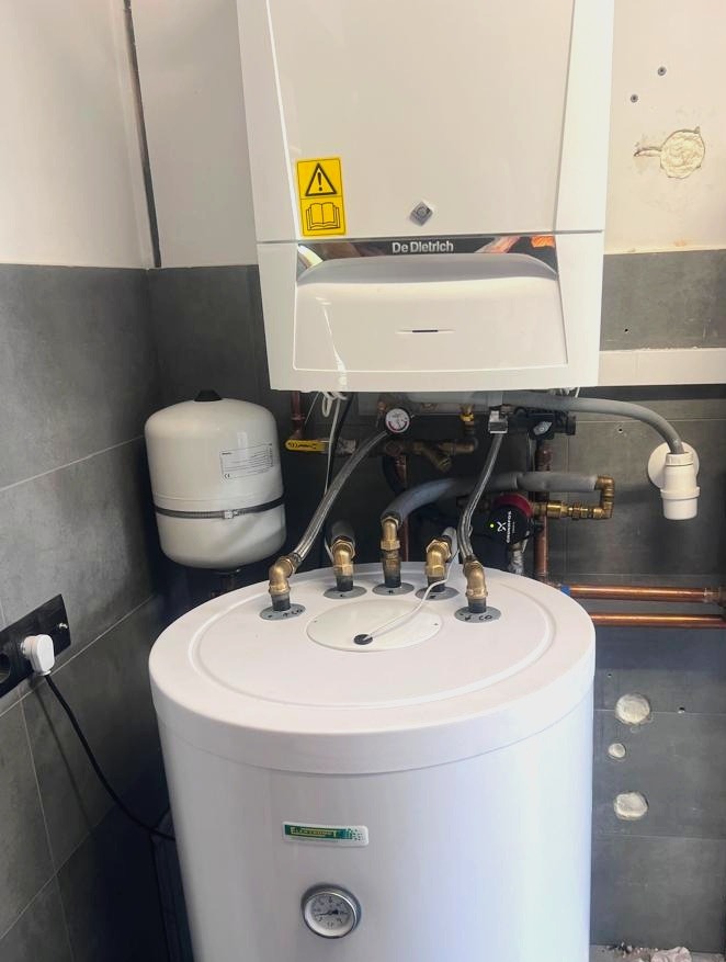 Biały piec gazowy De Dietrich zamontowany nad zasobnikiem ciepłej wody, widoczne podłączenia hydrauliczne i elektryczne, ściana z szarą glazurą.