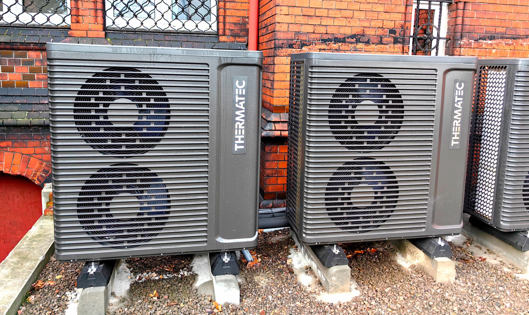 Dwa szare moduły pompy ciepła Thermatec ustawione na betonowych podstawach na tle ceglanego budynku z oknami z metalową kratą.