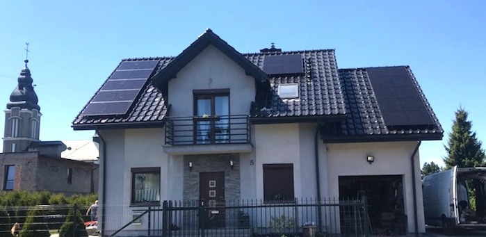Dwukondygnacyjny dom jednorodzinny z ciemną dachówką i panelami słonecznymi na dachu, widok z zewnątrz w słoneczny dzień; w tle wieża kościelna.