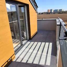 Lukesfera Szymczak Łukasz - Nowoczesny balkon z szarymi płytkami i metalową balustradą w słoneczny dzień. Cień balustrady tworzy geometryczne wzory na podłodze. Widok na otwartą przestrzeń.