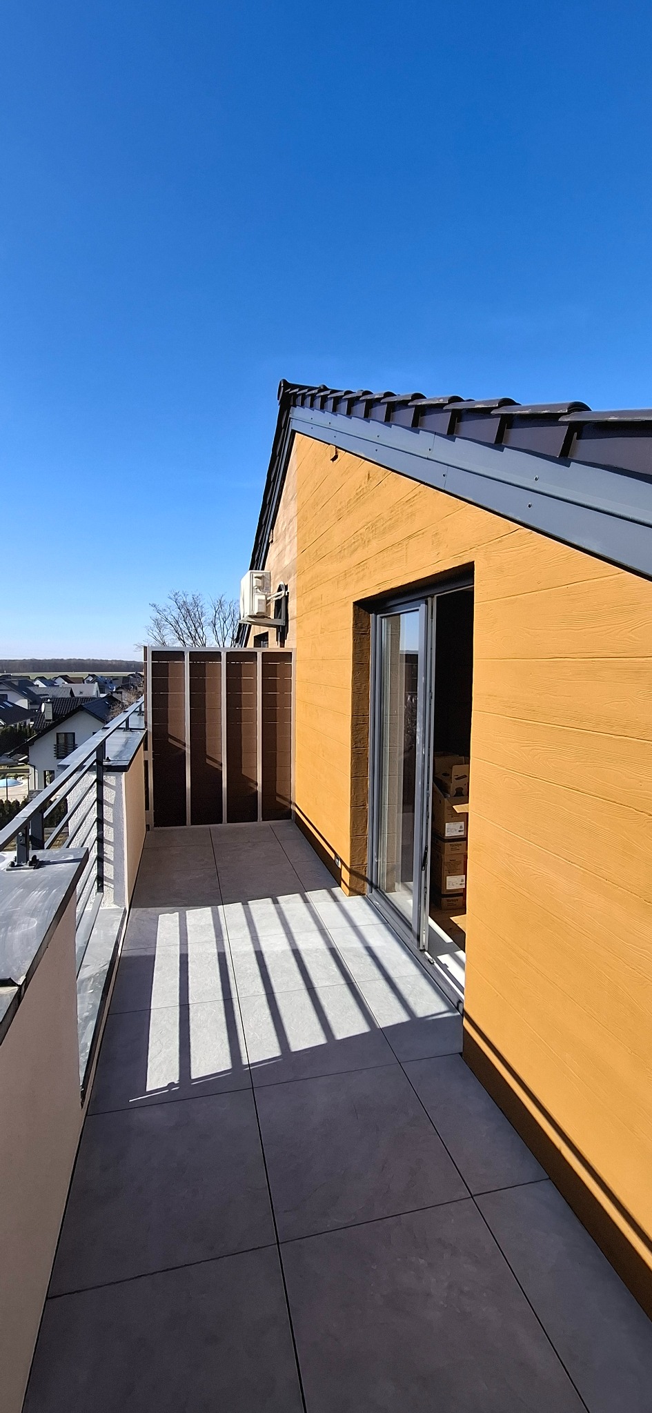 Nowoczesny balkon z szarymi płytkami i brązowymi panelami, oświetlony promieniami słońca tworzącymi cienie. Widok na okoliczne domy i błękitne niebo.