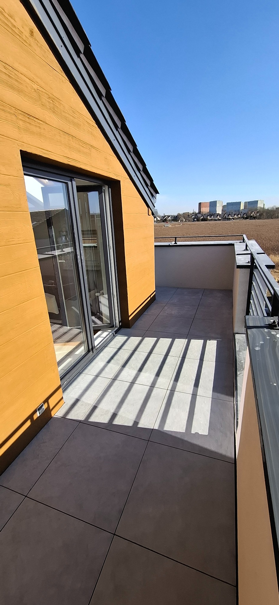 Nowoczesny balkon z szarymi płytkami i metalową balustradą w słoneczny dzień. Cień balustrady tworzy geometryczne wzory na podłodze. Widok na otwartą przestrzeń.