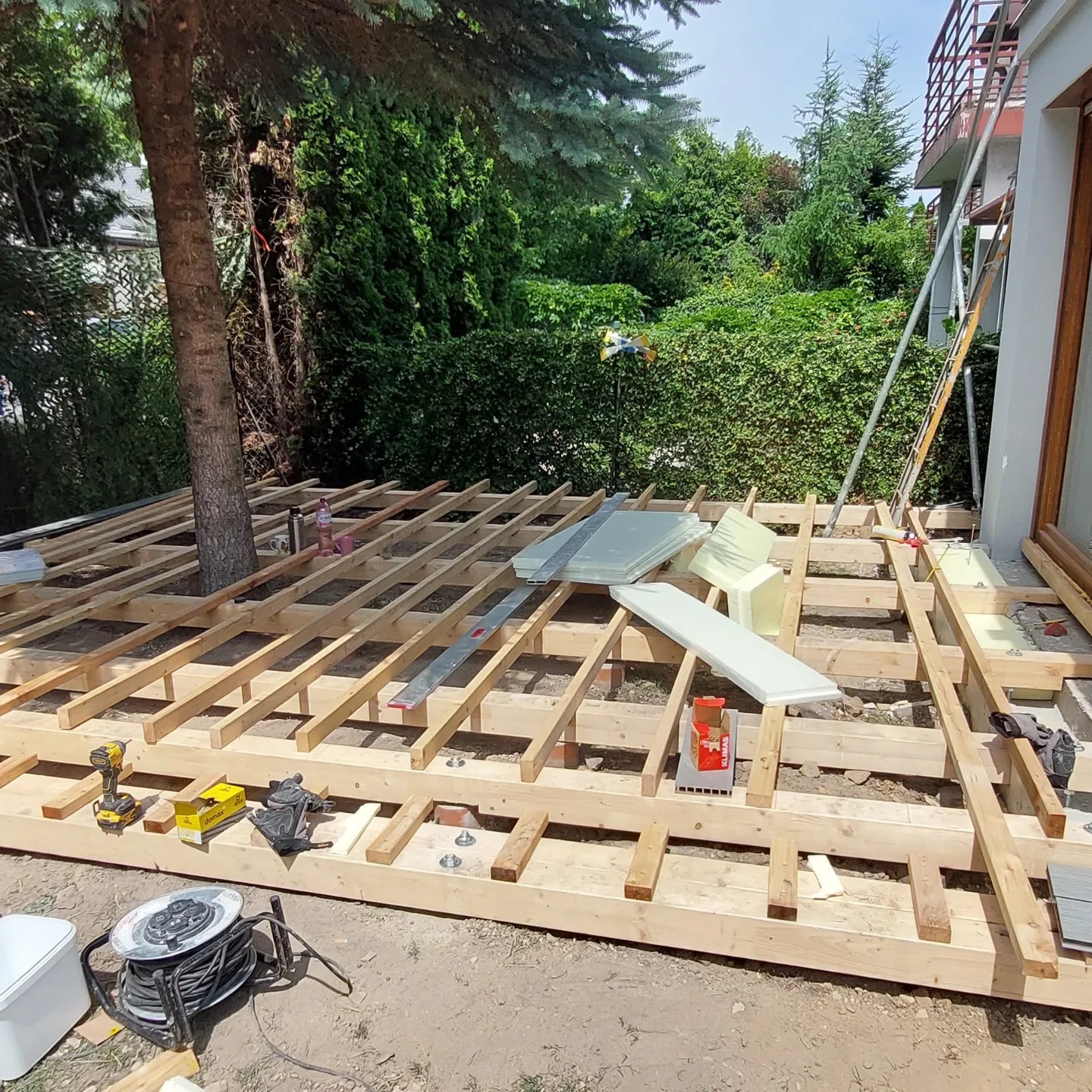 Montaż drewnianej konstrukcji tarasu na gruncie z drzewem, widoczne narzędzia i materiały izolacyjne. Drabina oparta o ścianę budynku. Częściowo ułożona podłoga.