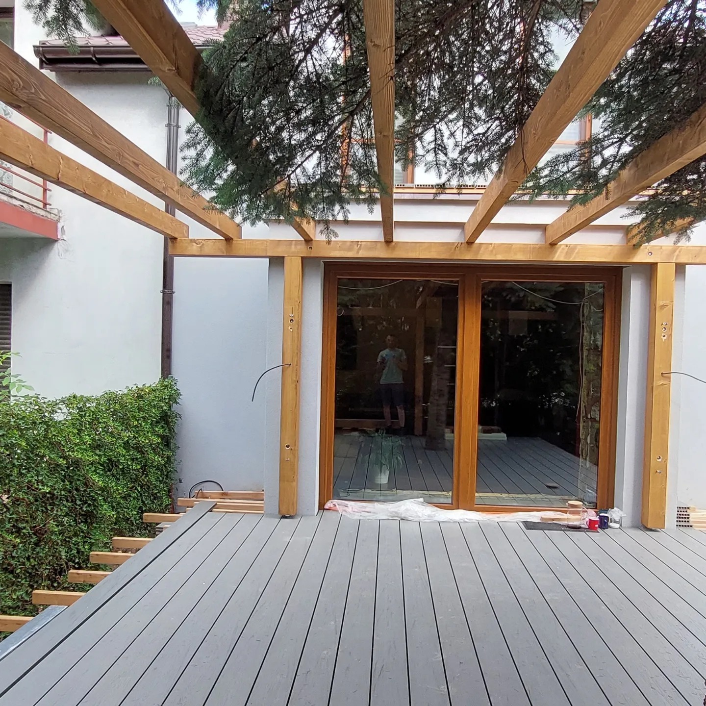 Montaż tarasu kompozytowego z drewnianą pergolą przylegającą do budynku. Widoczne deski tarasowe, konstrukcja pergoli i okno tarasowe z odbiciem osoby.