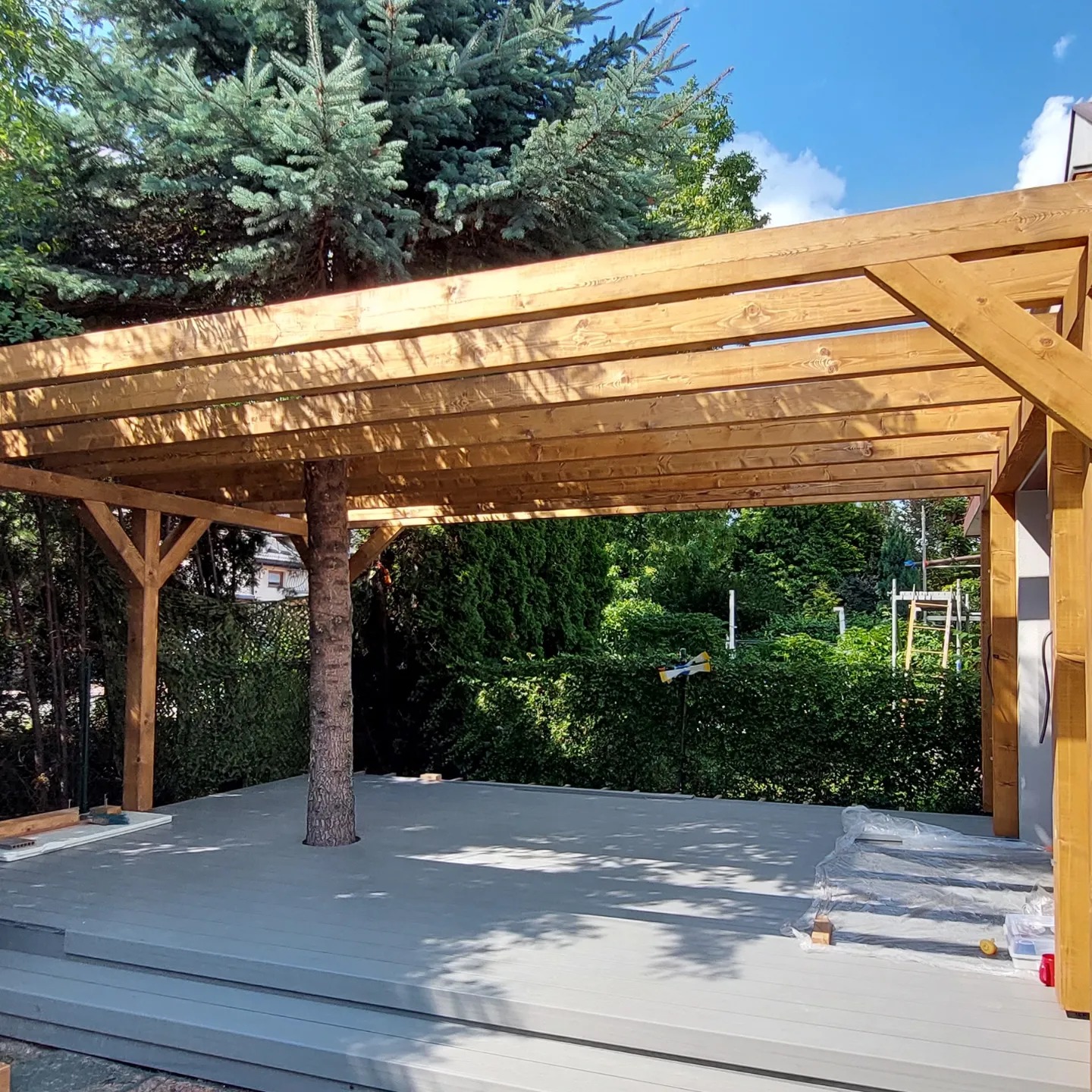 Drewniana pergola zadaszająca szary taras kompozytowy, zintegrowana z rosnącym drzewem. Jasne drewno konstrukcji, zielone otoczenie ogrodu i błękitne niebo w tle.