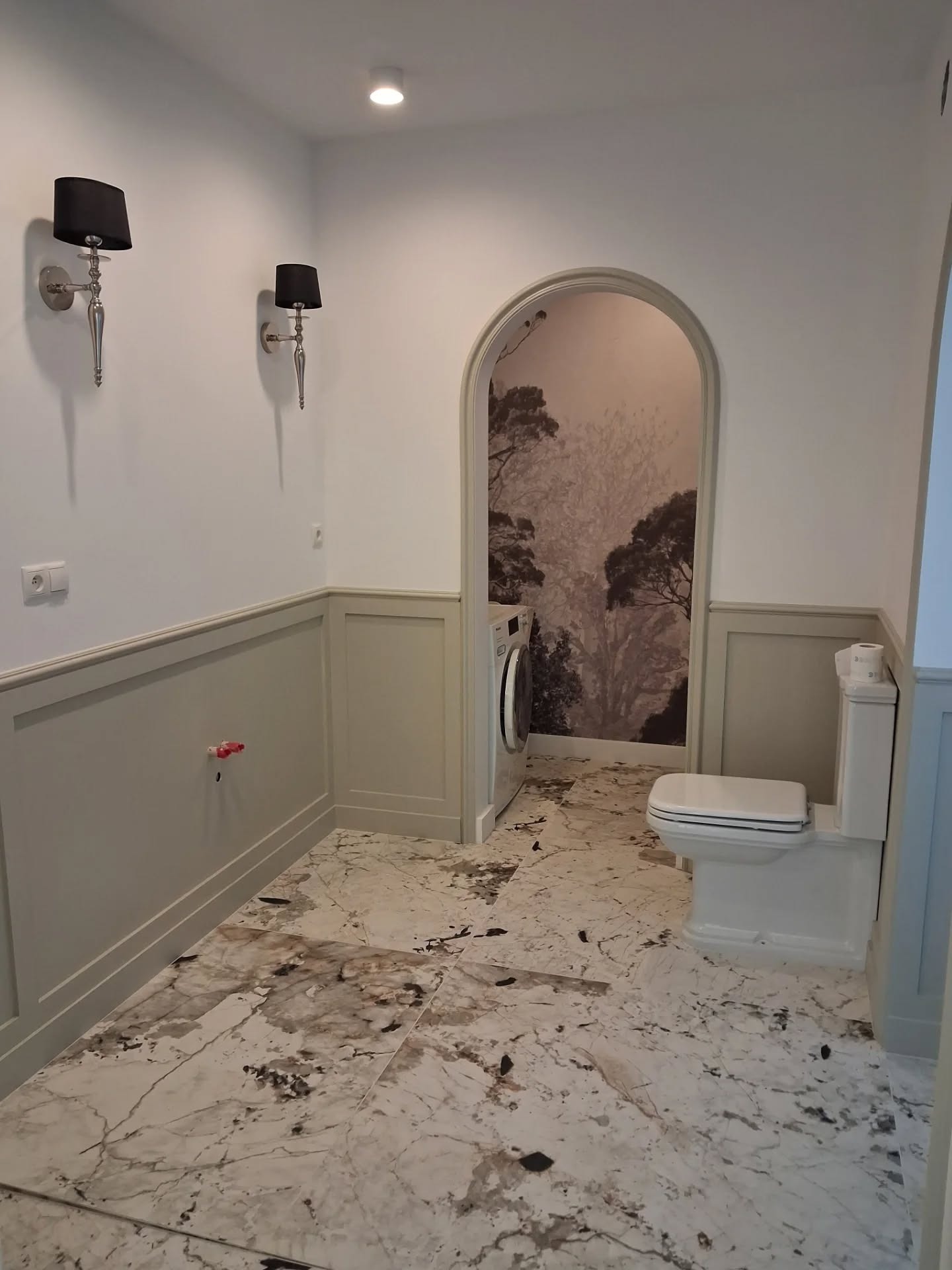 Elegancka łazienka po remoncie: marmurowe płytki podłogowe, ozdobne panele ścienne, designerskie kinkiety i wejście do pralni z fototapetą. Jasne, przestronne wnętrze.