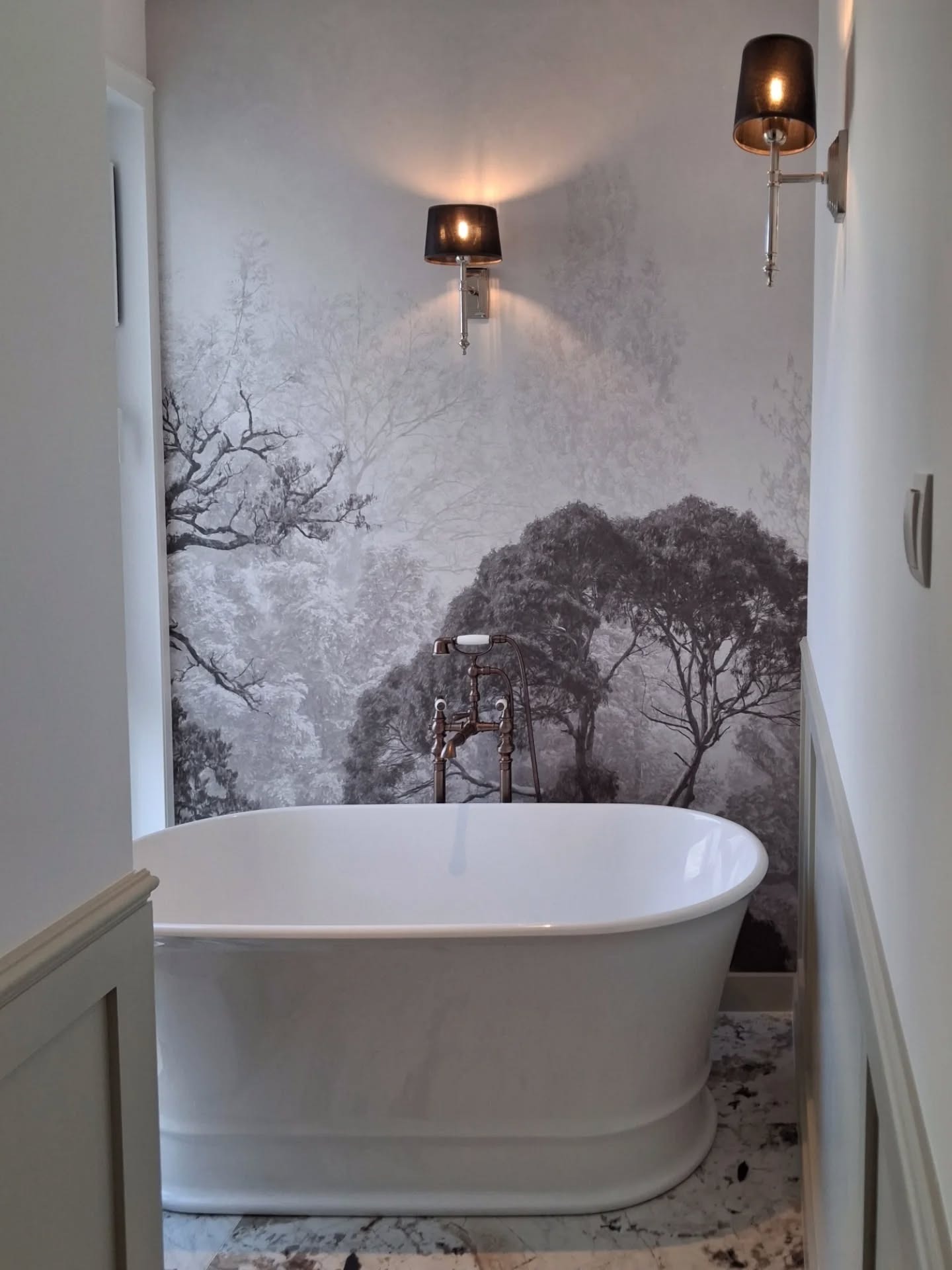 Elegancka łazienka z wolnostojącą wanną na marmurowej podłodze, ściana z fototapetą leśną i stylowe kinkiety z czarnymi abażurami. Nowoczesny design.