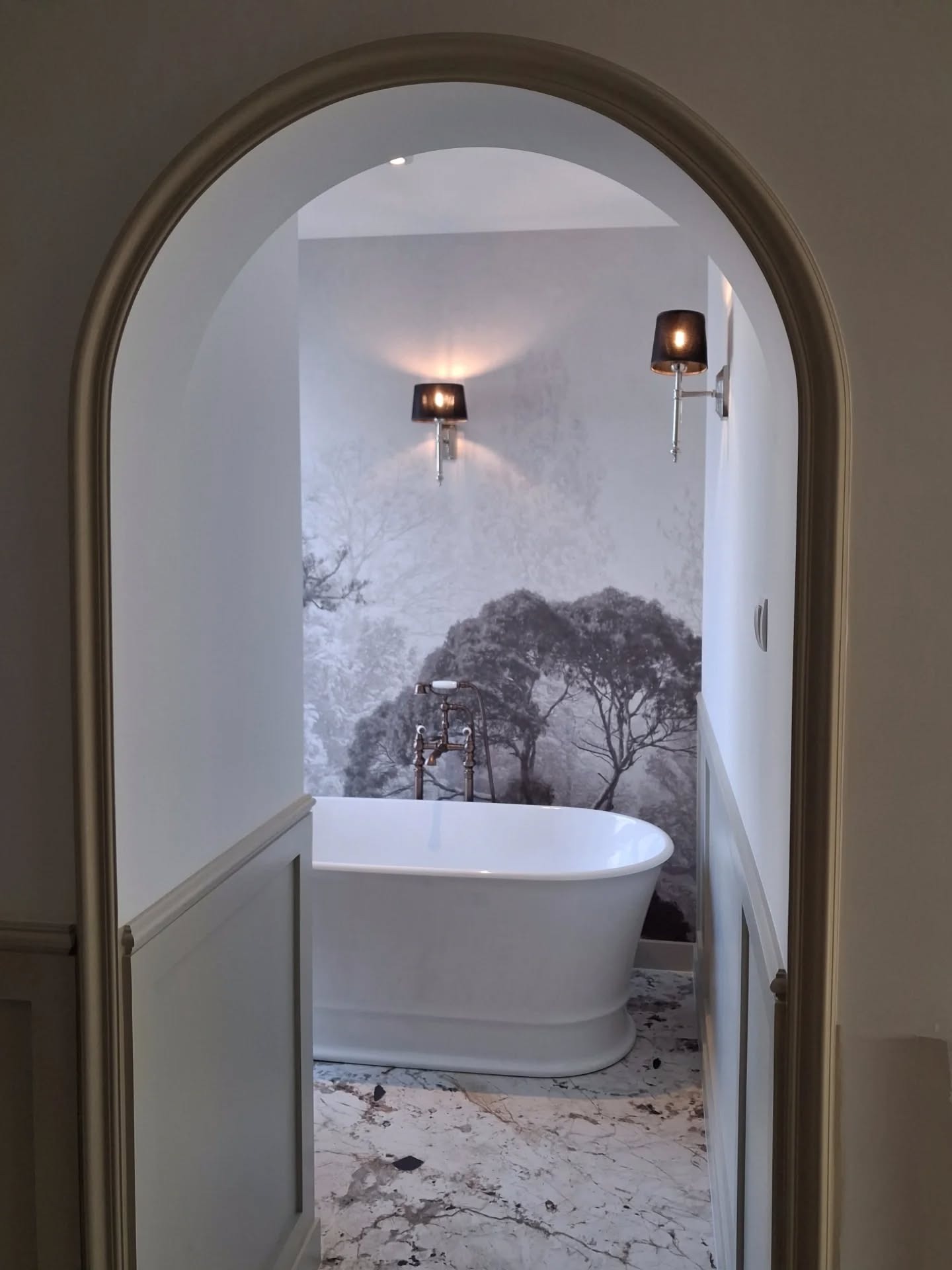 Elegancka łazienka z wolnostojącą wanną na marmurowej posadzce, oświetlona kinkietami z abażurami, widoczna przez łukowate przejście.