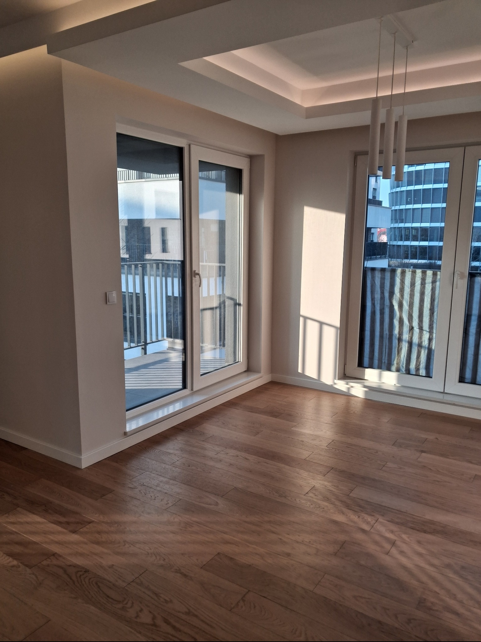 Jasne, przestronne wnętrze z parkietem w jodełkę, dużymi oknami balkonowymi i nowoczesnym oświetleniem sufitowym. Widok na budynek zza balkonu.