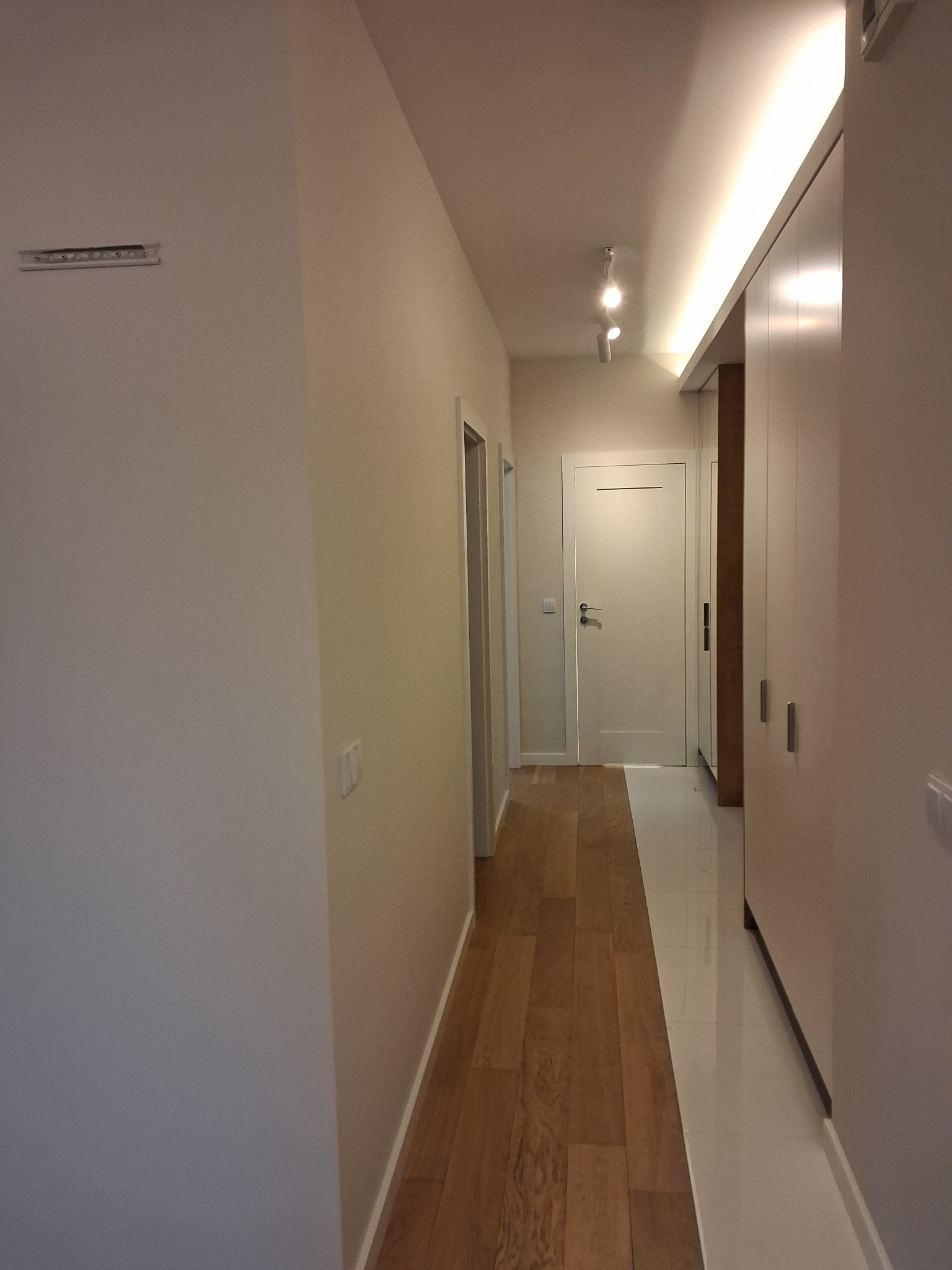 Nowoczesny korytarz z drewnianą podłogą przechodzącą w jasne płytki, oświetlenie punktowe i szafa w zabudowie w odcieniach bieli. Świeżo wykończone ściany w neutralnych barwach.