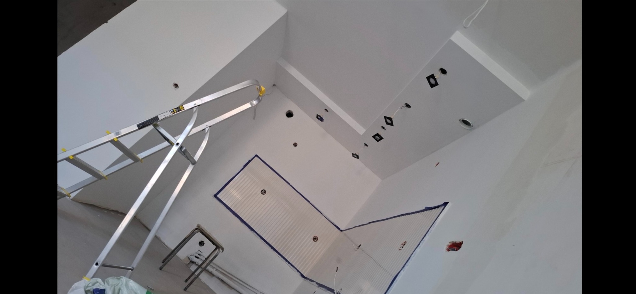 Białe wnętrze w trakcie malowania: drabina, zabezpieczone taśmą fragmenty ścian i sufitu, przygotowane otwory na oświetlenie. Widok z dołu, ukośny kadr.