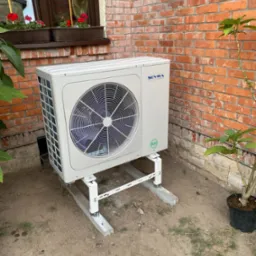 Biała jednostka zewnętrzna klimatyzacji zamontowana na metalowym stelażu przy ceglanym budynku z oknem i kwiatami w doniczkach, obok roślina w donicy.