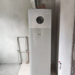 ThermTech Bartosz Wojdyło - Nowa, biała pompa ciepła Hisense, jeszcze w folii ochronnej, zainstalowana w surowym wnętrzu z widocznymi rurami i przygotowaną podłogą.