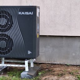 ThermTech Bartosz Wojdyło - Czarna pompa ciepła marki Kaisai zamontowana na białym stelażu przed elewacją budynku z widocznym otworem w fundamencie i butelką w środku.