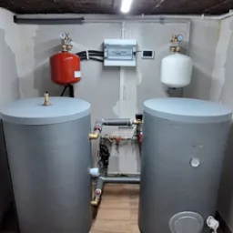 Dwa szare zbiorniki akumulacyjne połączone rurami w pomieszczeniu z drewnopodobną podłogą, widoczne również czerwony i biały zbiornik wyrównawczy, skrzynka elektryczna i surowy betonowy sufit z lampą.