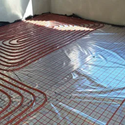 Czerwone rury ogrzewania podłogowego ułożone na folii z siatką w surowym wnętrzu, z blokami betonowymi w rogu i zwiniętymi rurami w tle.