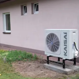 Biała pompa ciepła marki Kaisai zamontowana na betonowych fundamentach przy ścianie domu z trzema oknami, widoczny przewód w srebrnej izolacji.