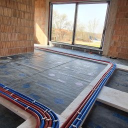 ThermTech Bartosz Wojdyło - Układanie rur ogrzewania podłogowego na izolacji w surowym wnętrzu. Czerwone i niebieskie rury przymocowane do styropianu. Widok na okno i ceglane ściany.