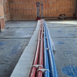 ThermTech Bartosz Wojdyło - Instalacja rur grzewczych (czerwone i niebieskie) na izolacji, w surowym wnętrzu z ceglanymi ścianami i oknem. Widok z perspektywy podłogi, rury ułożone równolegle.