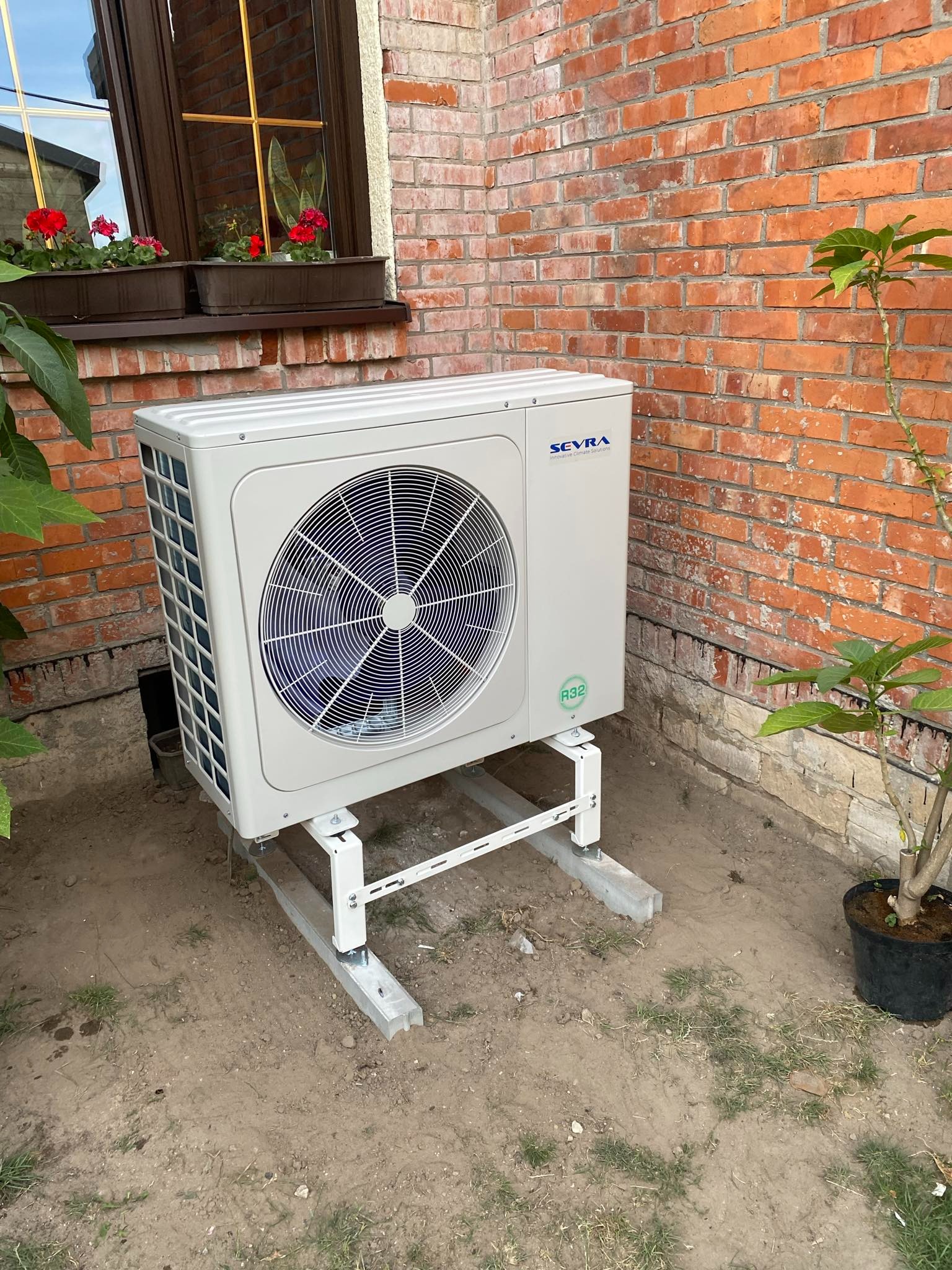 Biała jednostka zewnętrzna klimatyzacji zamontowana na metalowym stelażu przy ceglanym budynku z oknem i kwiatami w doniczkach, obok roślina w donicy.