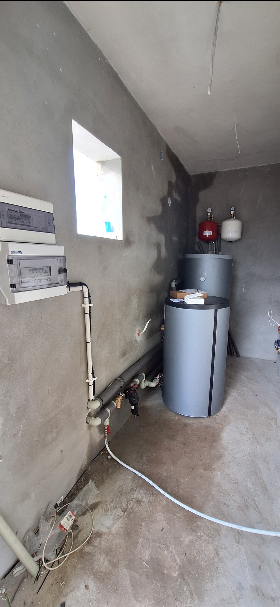 Dwa szare zasobniki ciepłej wody użytkowej z osprzętem hydraulicznym w surowym pomieszczeniu z betonowymi ścianami i podłogą, widoczne rury, zawory i skrzynki elektryczne.