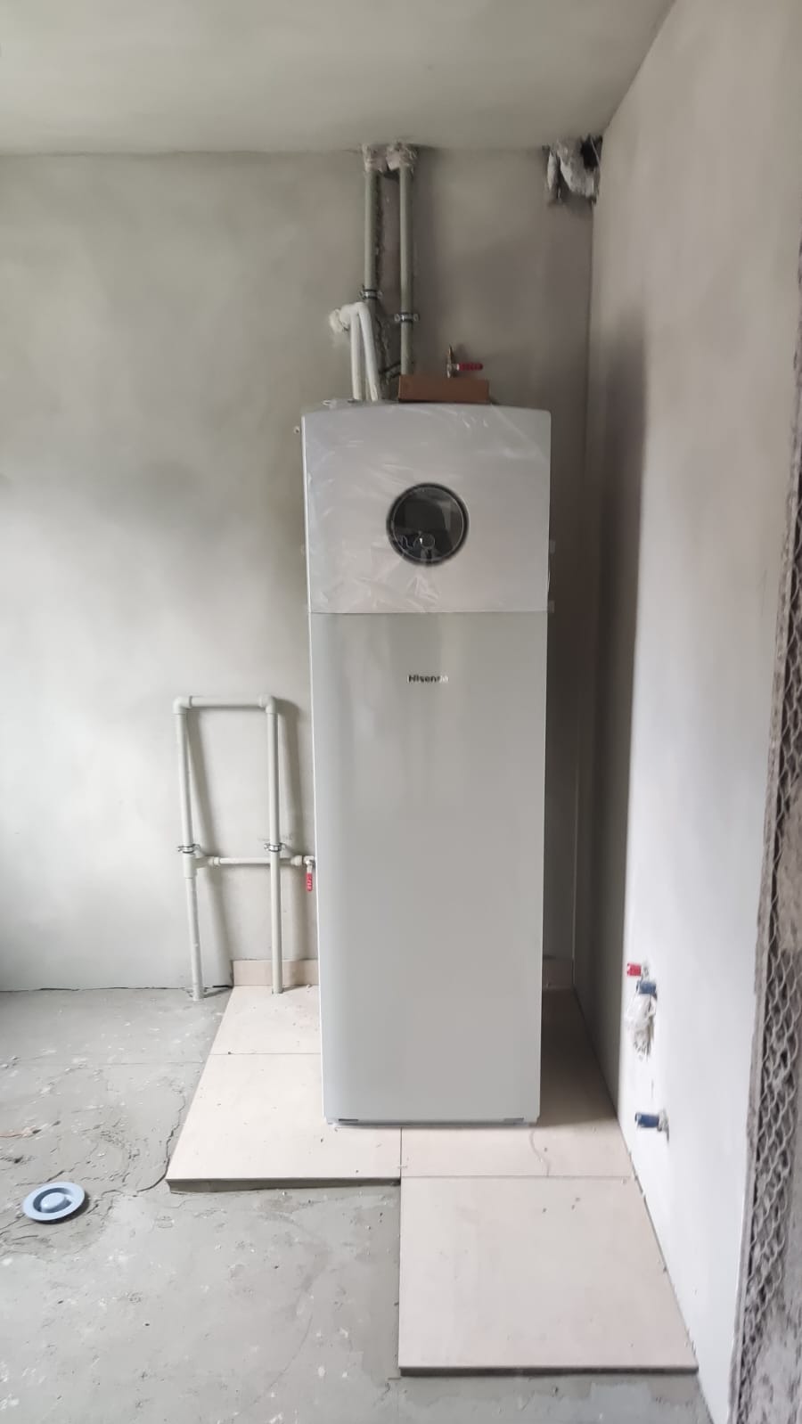 Nowa, biała pompa ciepła Hisense, jeszcze w folii ochronnej, zainstalowana w surowym wnętrzu z widocznymi rurami i przygotowaną podłogą.